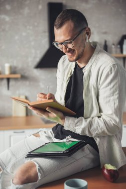 Elinde günlükle gülümseyen adam, elinde tablet olan bir not defteri, mutfakta eğleniyor. Çevrim içi eğitim.