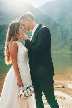Birbirlerine aşık romantik bir çift, gölün kıyısında öpüşüyorlar dağlar ve ışınların arka planına karşı. El ele tutuşmak. Yan görünüm