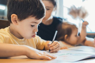 Genç bir çocuk evde annesiyle birlikte okul ödevini yapıyor, bir kitap yazıyor. Uzaktan eğitim. İlkokul ve evde eğitim, mesafe