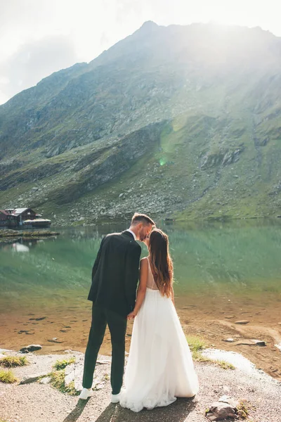 山を背景に湖の岸にキスを保持する若い新婚旅行のバックビュー かわいい山の風景 自然景観 — ストック写真
