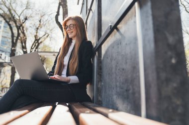Gözlüklü kızıl saçlı iş kadını dışarıda bankta otururken uzaktan bilgisayarla çalışıyor. Dijital girişimci ve kentsel yaşam tarzı kavramı.
