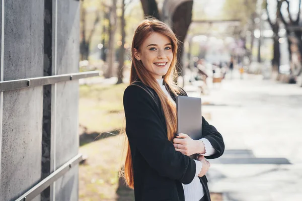 Happy redheaded businesswoman holding laptop and posing while walking on the city park. She uses laptop to communicate and work in Internet. Walk on autumn