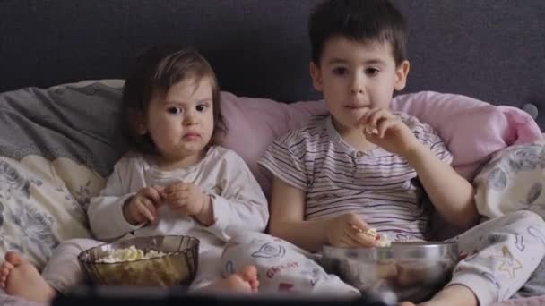 Δύο Παιδιά Βλέπουν Τηλεόραση Ξαπλωμένη Στο Κρεβάτι Τρώγοντας Ποπ Κορν — Αρχείο Βίντεο