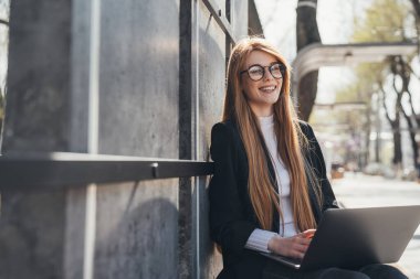 Gözlüklü bir kadın dizüstü bilgisayarda uzaktan çalışmaktan zevk alıyor, dışarıda bankta oturuyor. Modern iletişim. Çevrimiçi eğitim.