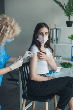 Tıbbi maske takan hemşire, beyaz kadın hasta tenini enjeksiyondan önce pamukla dezenfekte ediyor. Koronavirüs aşısı. Doktor.