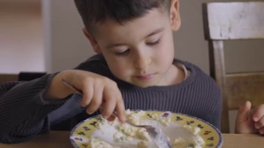 Küçük çocuk kaşığını tutuyor ve önünde bir kase yulaf lapası yiyor. Kahvaltıda yulaf lapası yiyor. Sağlıklı yiyecekler, diyet. Sağlıklı kahvaltı. Sağlıklı yemek..