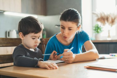 Anne bir masada oturuyor, oğluna ödevlerinde yardım ediyor, not defterine notlar yazıyor. Covid-19 sırasında evde uzaktan eğitim, çevrimiçi eğitim