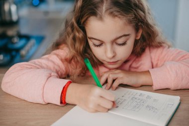 Caucasian young schoolgirl sitting at table, writing in her notebook or diary. Front view. Internet technology. Distance education. Online education.