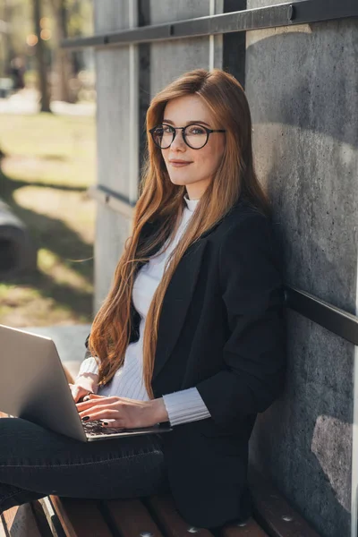 Yaz parkında dizüstü bilgisayarda çalışan bir kadın bankta oturuyor, gülümsüyor ve kameraya bakıyor. Serbest çalışma konsepti. Çevrimiçi eğitim, uzaktan çalışma ve