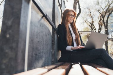 Red-headed young woman sitting in public park chatting over laptop computer sitting on a wooden bench. Using laptop and mobile phone. Distance learning online