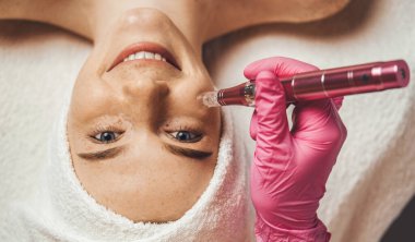 Cosmetologists hand doing procedure of microneedling to the patients smiling face. Aesthetic cosmetology, face care. Fractional Needle mesotherapy with clipart