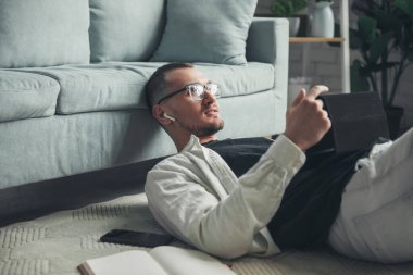 Portrait of a relaxed caucasian man lying on the floor leaning his head on the sofa speak on webcam video call on tablet. Home office. Using tablet, watching.