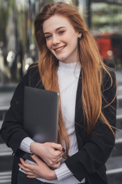 Front view smiling redhead woman holding laptop pc computer standing outdoors in front of office building. Modern beauty portrait. Young lady is working.