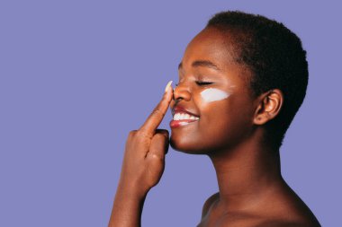 Afro woman applying cream on her cheeks and with her index finger spreads the cream on her nose isolated over purple background with blank space for text. Side