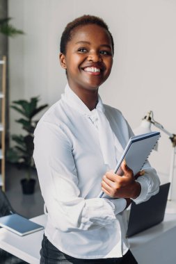 Closeup portrait of a beautiful african american woman in white shirt posing holding notebook, standing in the office. Attractive beautiful girl. Beautiful girl