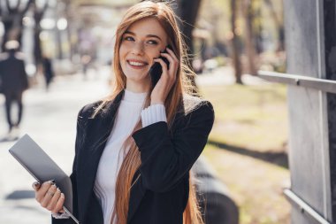 Smiling young caucasian red-haired businesswoman talking on mobile phone, outdoors in the park. Phone communication. Business call.