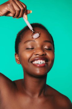 Smiling afro woman massaging face skin with jade roller isolated over green background. Beautiful girl portrait. Attractive beautiful girl. Body care.