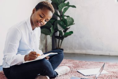 Afro student girl sitting on floor preparing homework for tomorrows lesson at the university. Course, book, speaking on video call on laptop, writing notes.