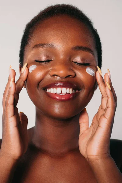 Portrait of black woman applying face cream on her cheeks, smiling at camera, isolated against white backdrop. Beauty treatment. Beautiful girl portrait.