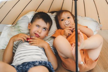 The older brother makes unpleasant faces because he has to sit with his younger sister lying on the floor under an umbrella at home. Beautiful colorful