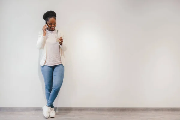 African american woman talking on mobile phone with friend standing with glass of coffee against white wall. Drinks caffeine beverage. Using modern technologies