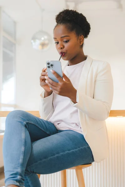 Portrait of an african american woman texting her friends on smart phone, sitting at a coffee shop, drinking a beverage. Internet technology. Beauty portrait.