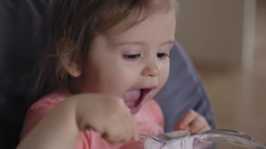 A baby girl sitting in high chair eating yoghurt with spoon from her big bowl. Portrait for lifestyle design. Healthy meal.