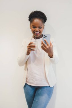 Smiling afro american woman doing a video call on mobile phone, drinking a glass of beverage, isolated over white background. Creative new idea and selfie.