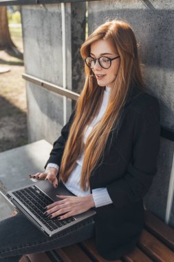 Smiling businesswoman using her laptop while sitting on the bench in the park and working online. Business, education, lifestyle concept. Happy teenager