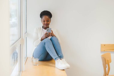 African woman sitting on the windowsill of a cafe, writing messages to her customers. Checking email. Electronic wireless device concept