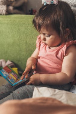 Baby girl child holding a a fairy tale book looking at illustration sitting on the couch. Beautiful girl portrait. Family day concept. Happy family, childhood