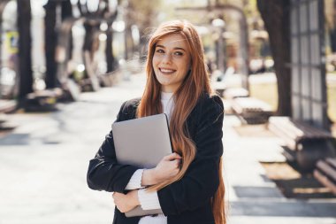 Smiling ginger woman looking away, carries laptop waits for someone in the park. Caucasian woman working on laptop, freelance