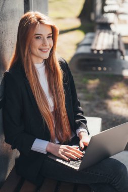 Red-headed woman working with laptop outdoor in the park, smiling while posing looking at camera. Internet technology. Office worker.