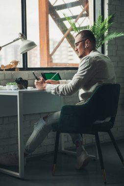 Caucasian man holding a phone in one hand, pencil in other hand using tablet, sitting on chair in the office. Concept of job search online. Businessman using
