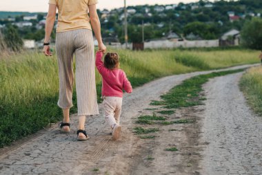 Back view of a mother walking holding hands with her little baby on countryside road. Parent, child. Beautiful girl portrait. Baby development. Back view