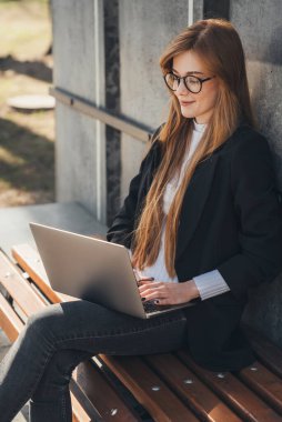 A red-headed girl working with a laptop on a bench in the park. The concept of an open-air office. Fashion model. Travel fashion. Attractive beautiful girl.