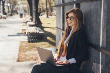 Smiling business woman working on laptop in the public place on bench. Technology concept, tablet, notebook, teleworking, telecommuting, zenithal.