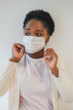 Black female with curly hair putting on medical mask while standing in a room with closed air, and looking away. Isolated over white wall. Respiratory virus