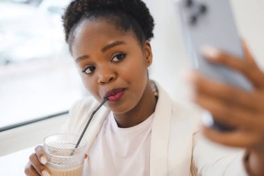 Young happy and attractive black afro woman sitting in a coffee shop taking a selfie on cell phone. Making selfie via application on portable. Beauty portrait.