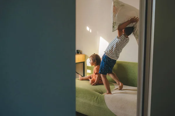 View from behind the bedroom door, the brother is holding a pillow in his hand, wanting to surprise his baby sister who is sitting on the couch, to start a