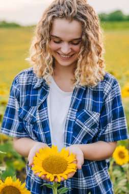 Curly woman farmer standing in the middle of a sunflower field, holding a sunflower in hand and looking at it with admiration. Agricultural worker. Summer