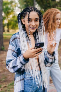 Young girl with afro pigtails holding phone showing v sign while looking at camera. Beautiful tiktok vloggers enjoying free time in the park