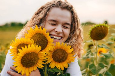 Happy curly girl embracing sunflower flowers in a field of sunflowers at sunset. Beauty portrait. Natural summer landscape. Agronomist and farmer inspect