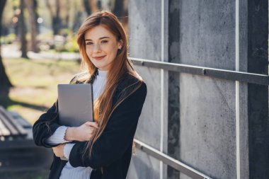 Smiling ginger woman holding laptop while walking outdoors through the city park on sunny day. Gorgeous redhead businesswoman. Beauty portrait. Business