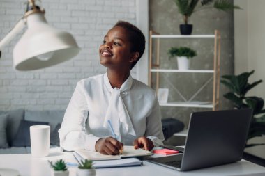 Smiling african-american woman with curly hair working remotely using laptop computer, write in notebook. Smart intelligent woman entrepreneur sitting at the
