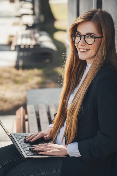 Read-headed student woman in black jacket sitting on bench in park outdoors, looking and smiling at camera use laptop. People urban lifestyle concept