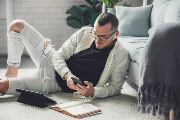 Man in eyeglasses with mobile phone studying from home lying on the floor in a modern and stylish interior. Education internet technology. Distance education