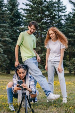 Three gorgeous bloggers recording video on mobile phone in the city park to attract followers. Share content. App post.