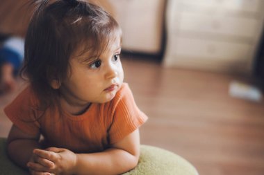 Pensive little daughter looking attentively and thoughtfully away, while she is leaning on the sofa with her arms. Portrait baby girl. Carefree childhood. Skin