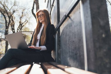 Side view ginger woman student wearing eyeglasses studying online outdoors in the park, using laptop. Freelance, e-learning.